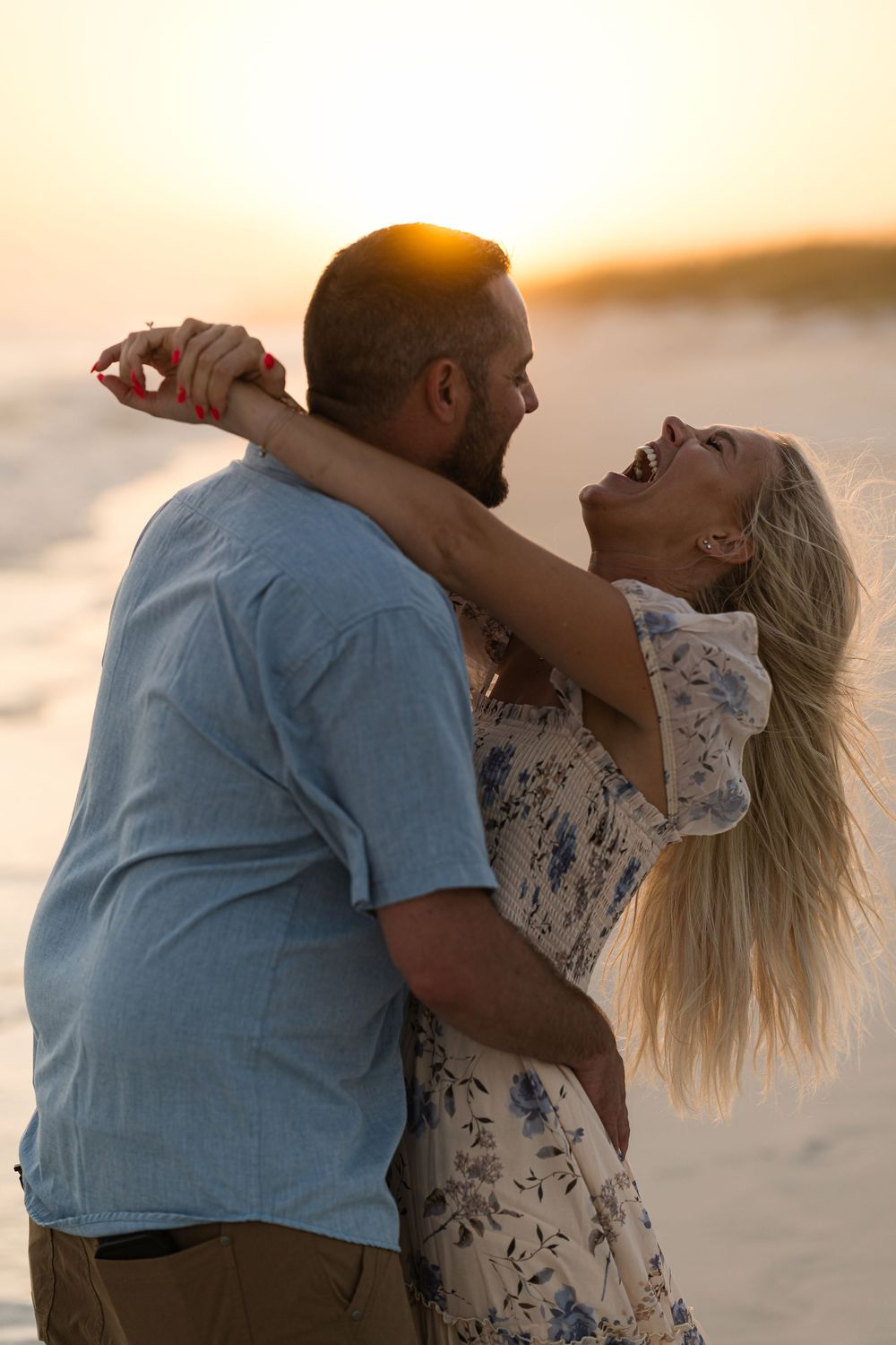 Couple laughing and embracing on a beach at sunset, woman in floral dress, man in blue shirt.