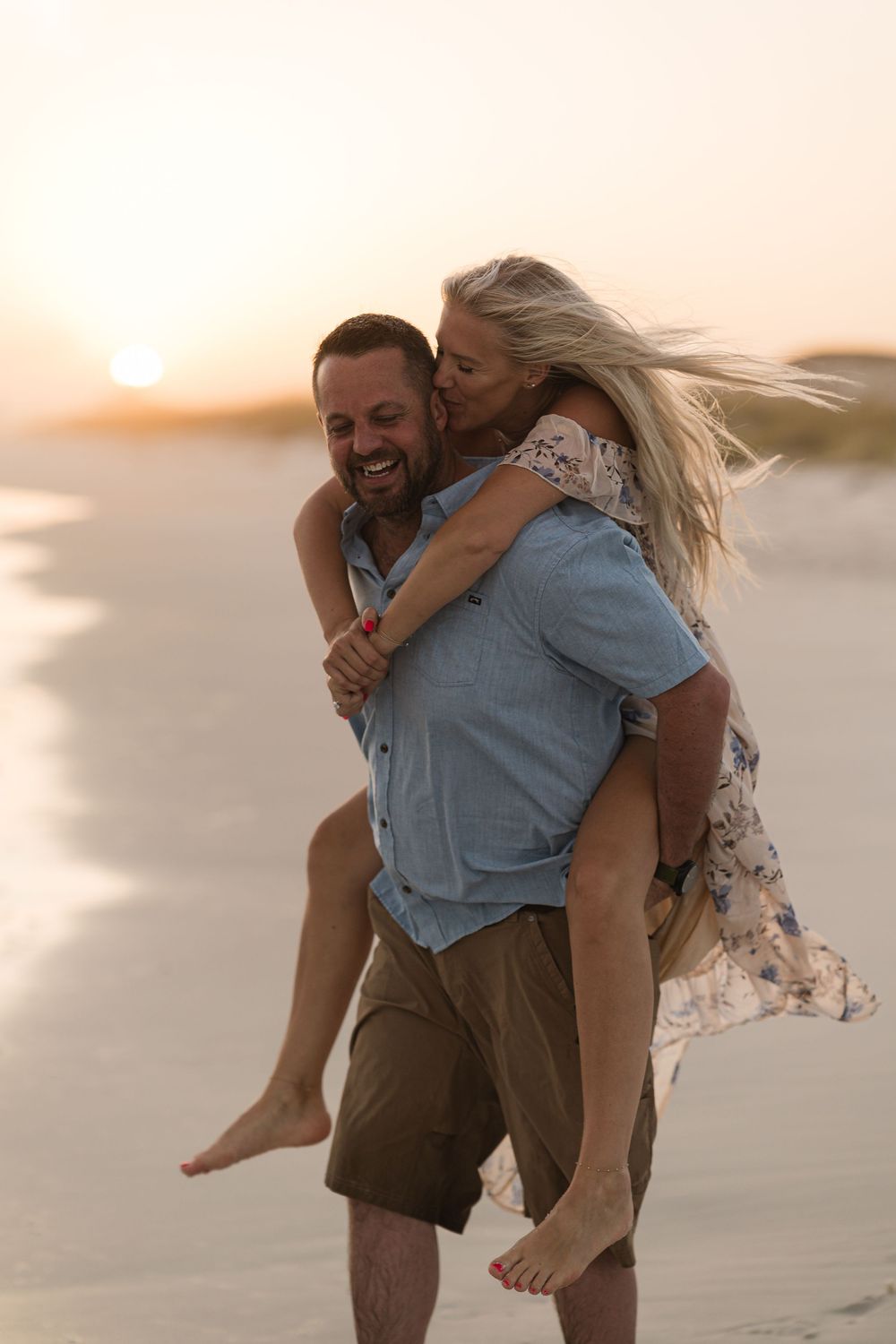 Couple enjoying a piggyback ride on the beach at sunset, woman kissing man's cheek while he laughs.