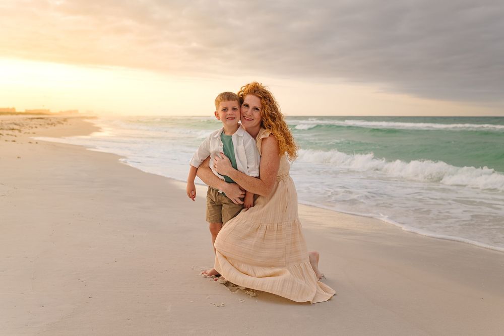 Woman in flowing dress kneeling on sandy beach hugging young boy at sunset with ocean waves behind them.