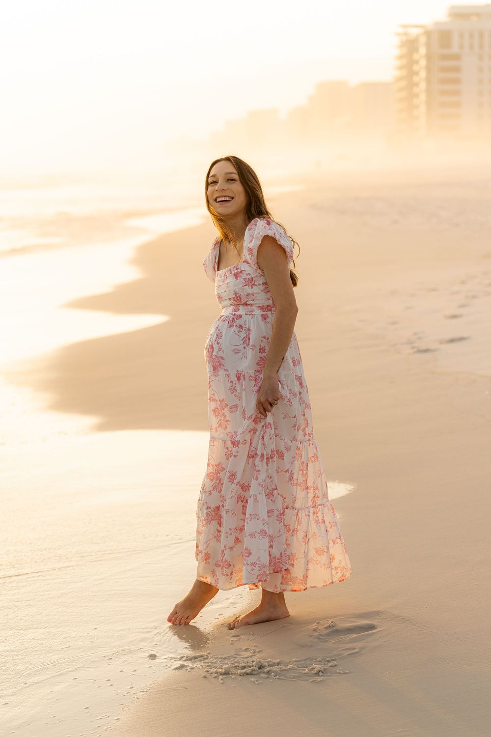 Smiling pregnant woman in floral maxi dress walking barefoot on sandy beach at golden sunset.