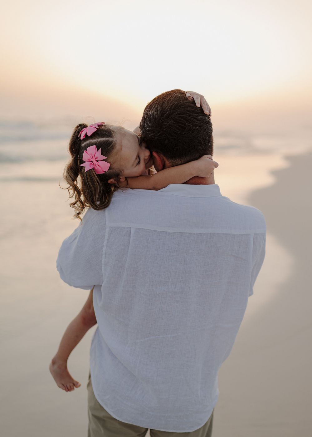 A father holds his young daughter with pink hair bows on a beach at sunset.