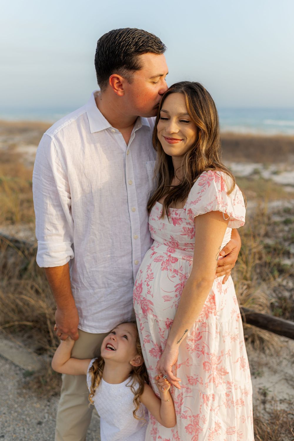 Pregnant woman in floral dress with husband kissing her cheek and young daughter laughing at beach.