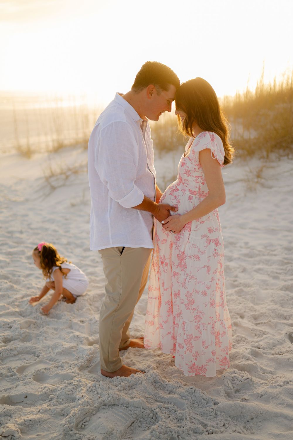 Pregnant couple sharing a tender moment on sandy beach at golden sunset, with young child playing in sand nearby.