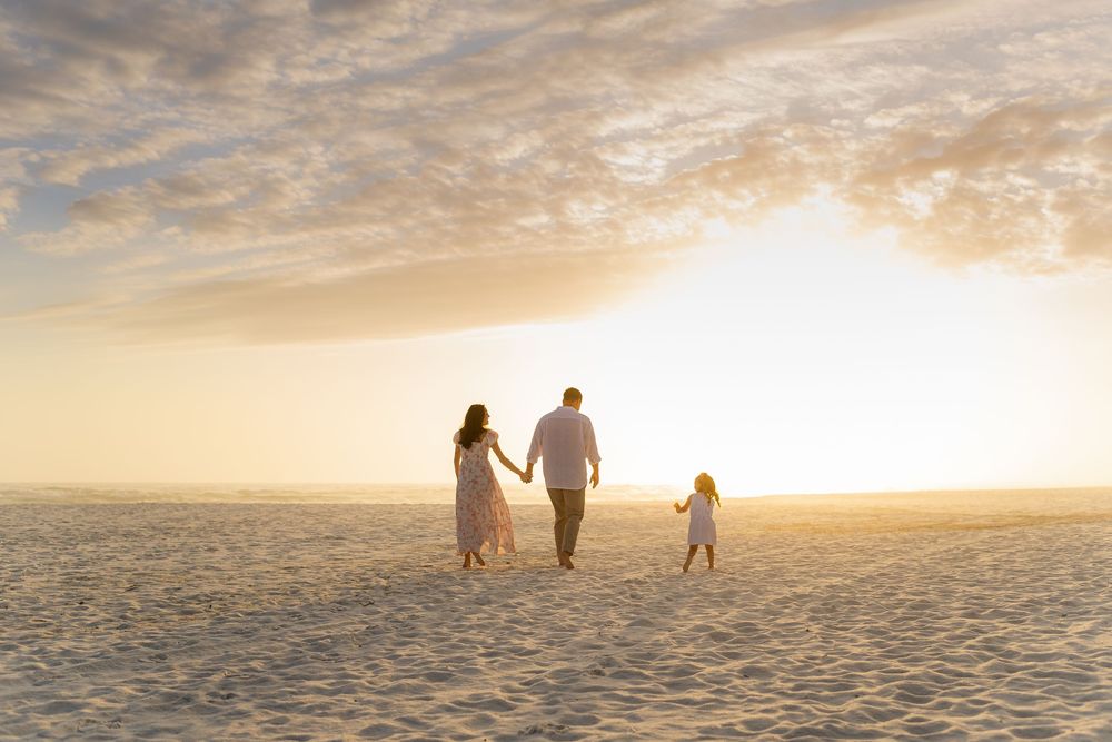 Family of three walking hand-in-hand on a sandy beach at golden sunset, rear view with warm glowing sky.