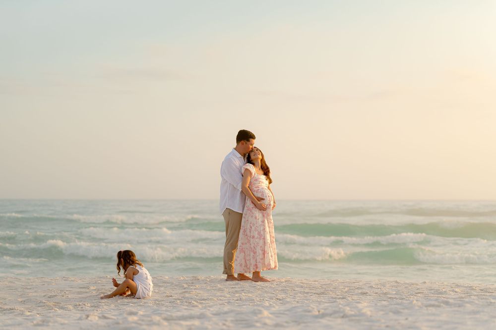Couple embracing on a sandy beach at sunset while a young child plays nearby, with ocean waves in the background.