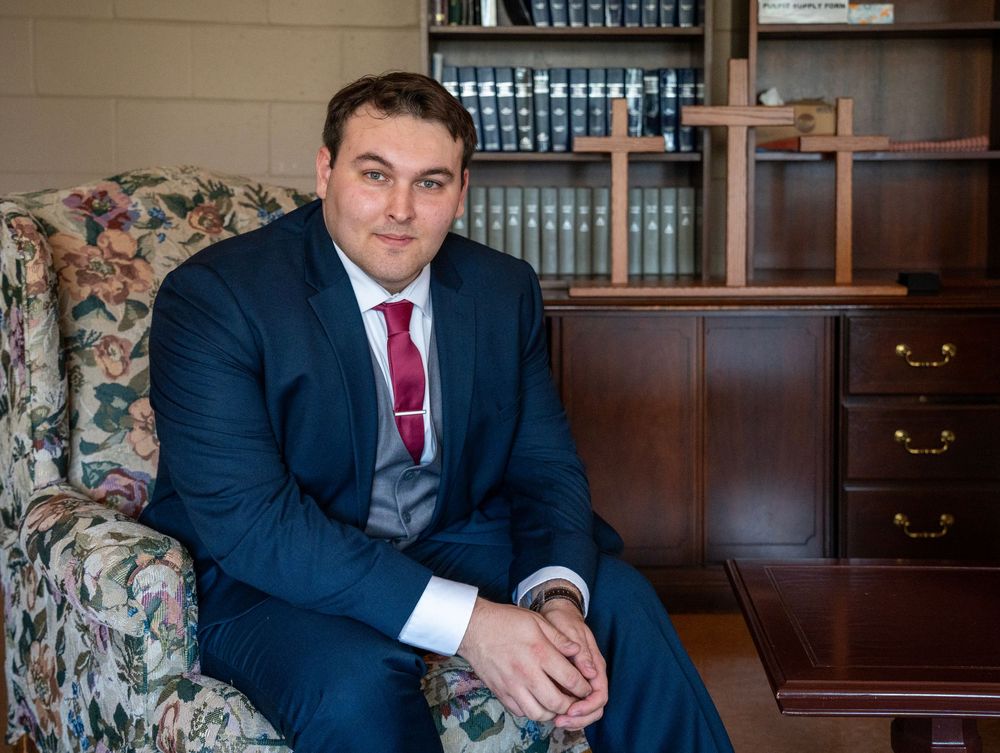 Groom portrait sitting on a floral chair looking at the camera with a bookshelf in the background.
