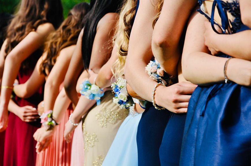 Row of formal dresses at prom or wedding with corsages and flowers visible on wrists against blurred background.