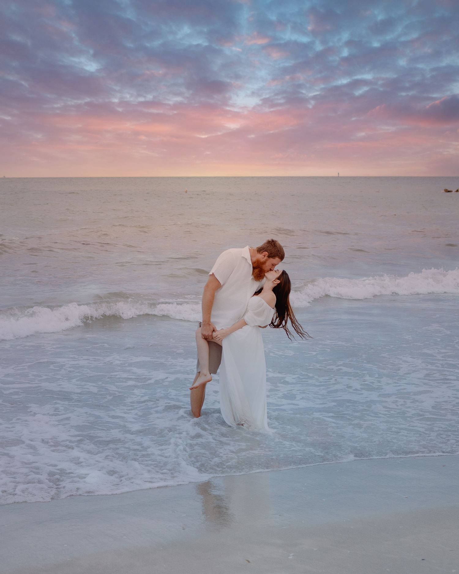 A couple sharing a romantic moment in white attire at the beach during sunset with pink and purple clouds above.