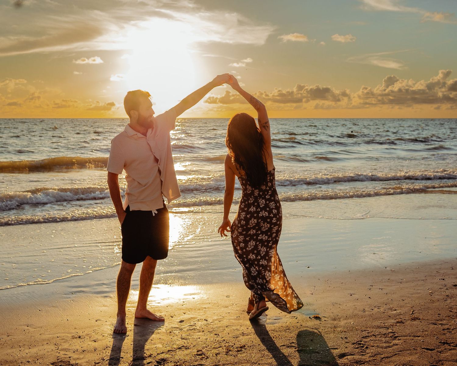 A couple creates a heart shape with their arms while dancing on a beach during golden hour sunset.