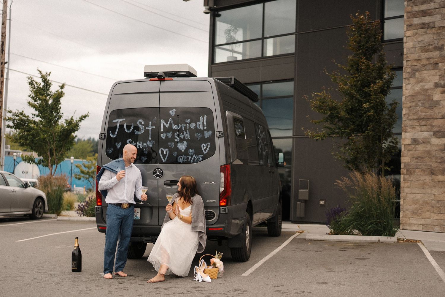 Two people stand next to a gray van in a parking lot with drinks and shopping bags beside them.