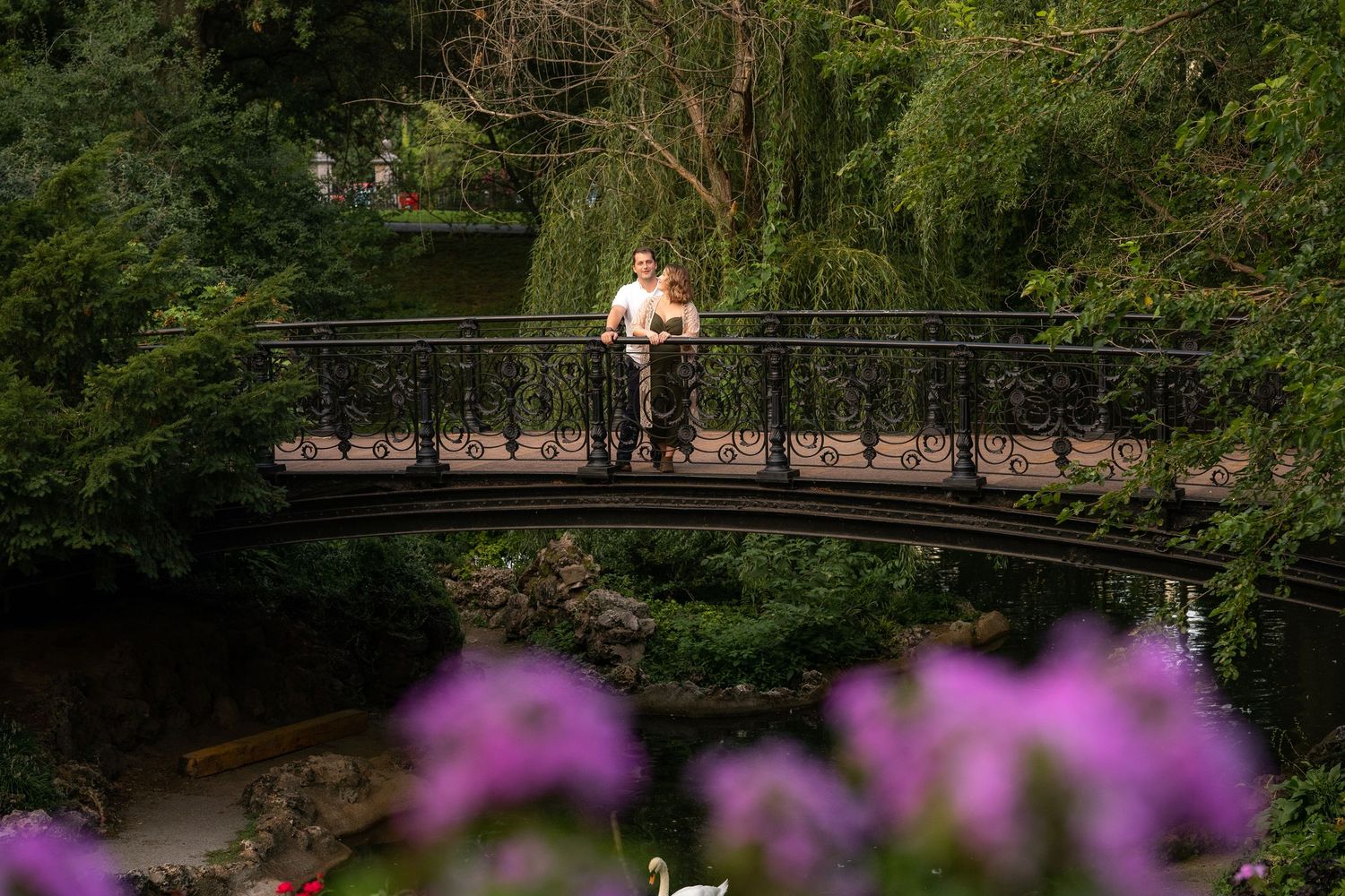 Portrait of a couple standing on a iron bridge with greenery in the background and purple flowers blurred out in the foreground.