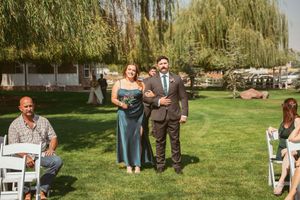 Wedding party members walk down the grassy aisle lined with white chairs under weeping willow trees.