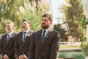Wedding party members in suits and formal attire standing in line outdoors with trees in background.