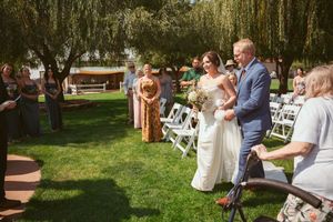 A wedding ceremony takes place outdoors on a bright sunny day with white chairs and willow trees in the background.