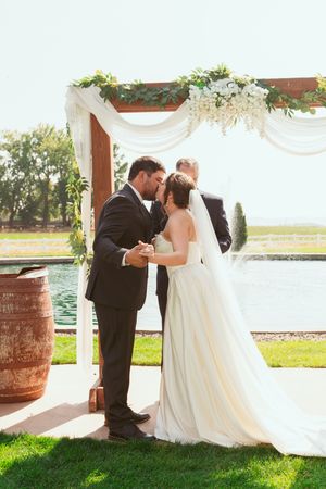 Wedding couple shares an intimate moment under a draped wooden arch decorated with white flowers beside a pond.