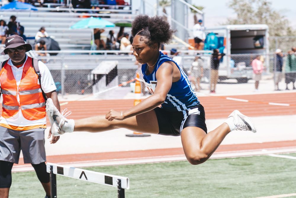 Track athlete in blue uniform clearing hurdle during competition.