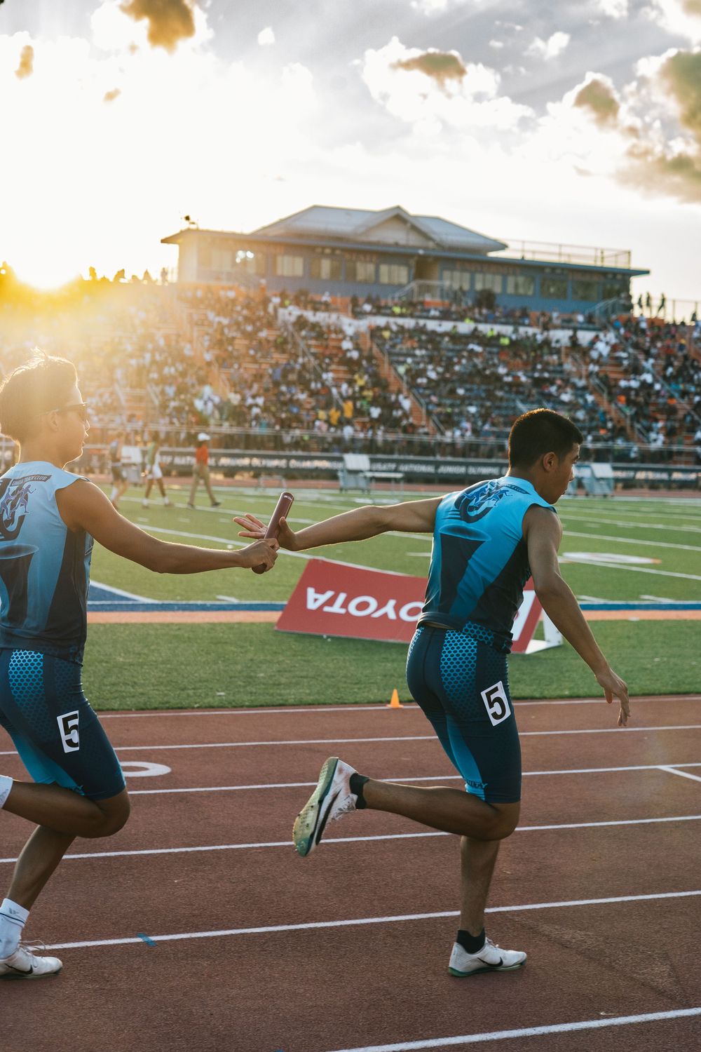 Relay team exchanging baton during sunset track meet.