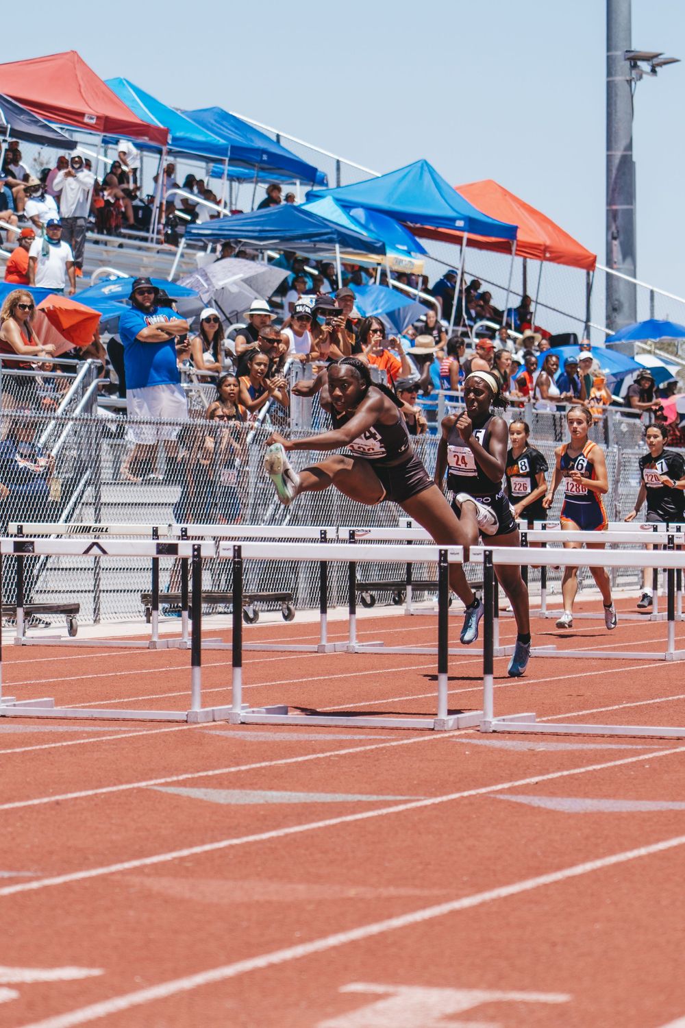 Athletes competing in hurdles race with crowd and tents visible in background.