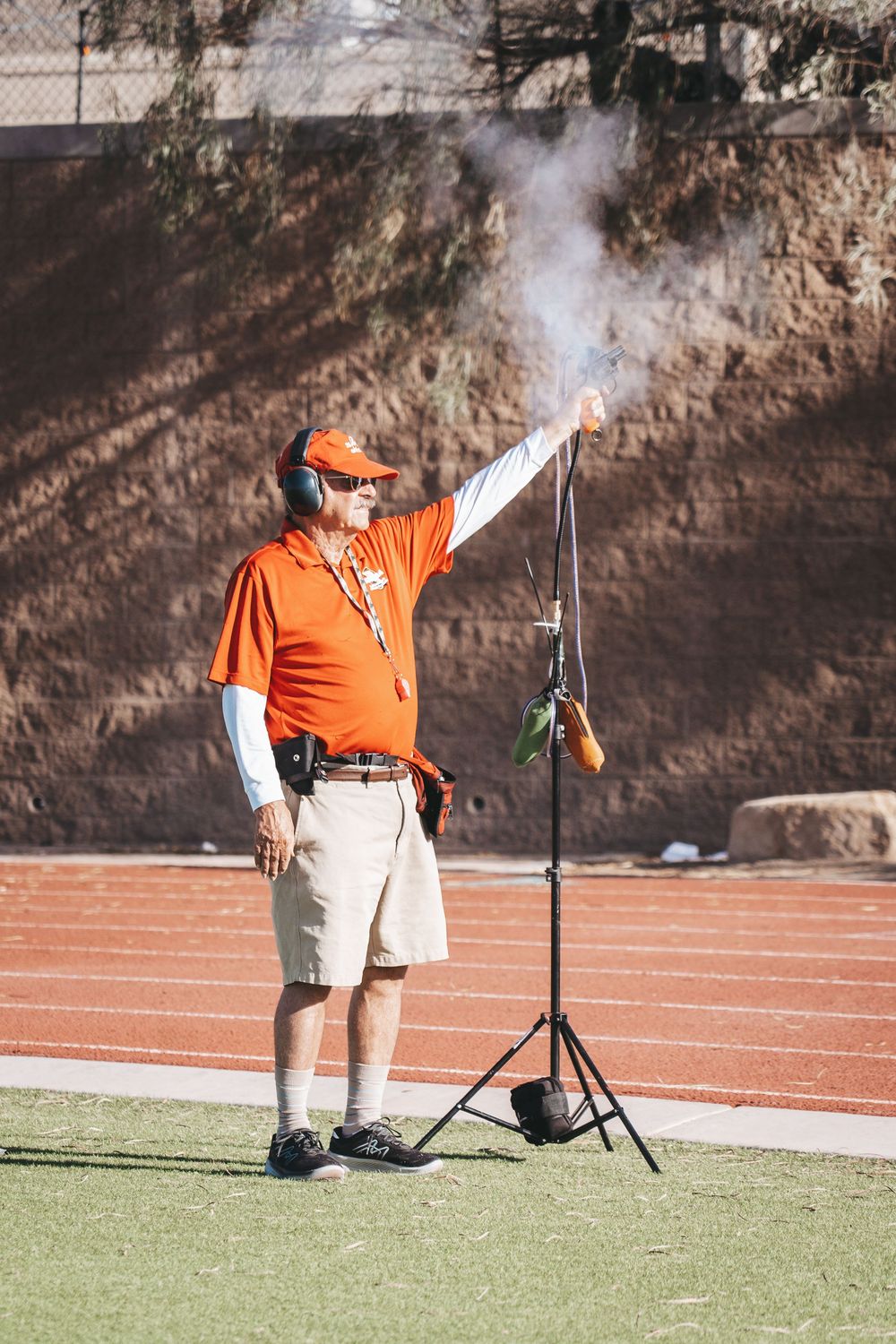 Official in orange shirt operates starting pistol at track meet on sunny day.