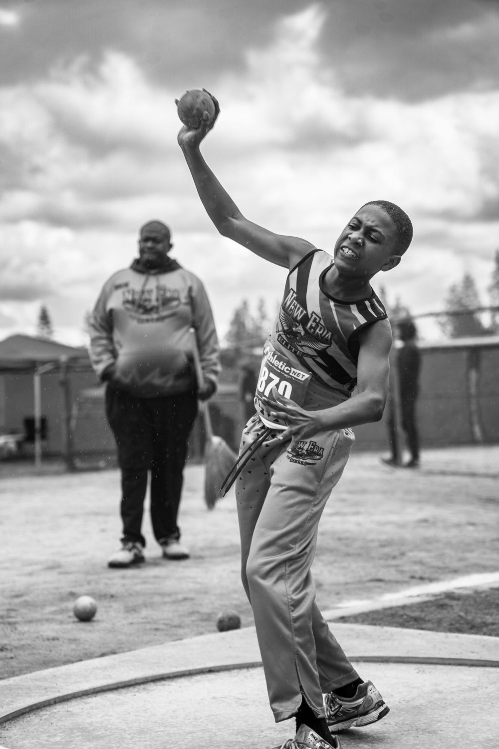 Young athlete throws shot put at outdoor track meet in dramatic black and white photo.