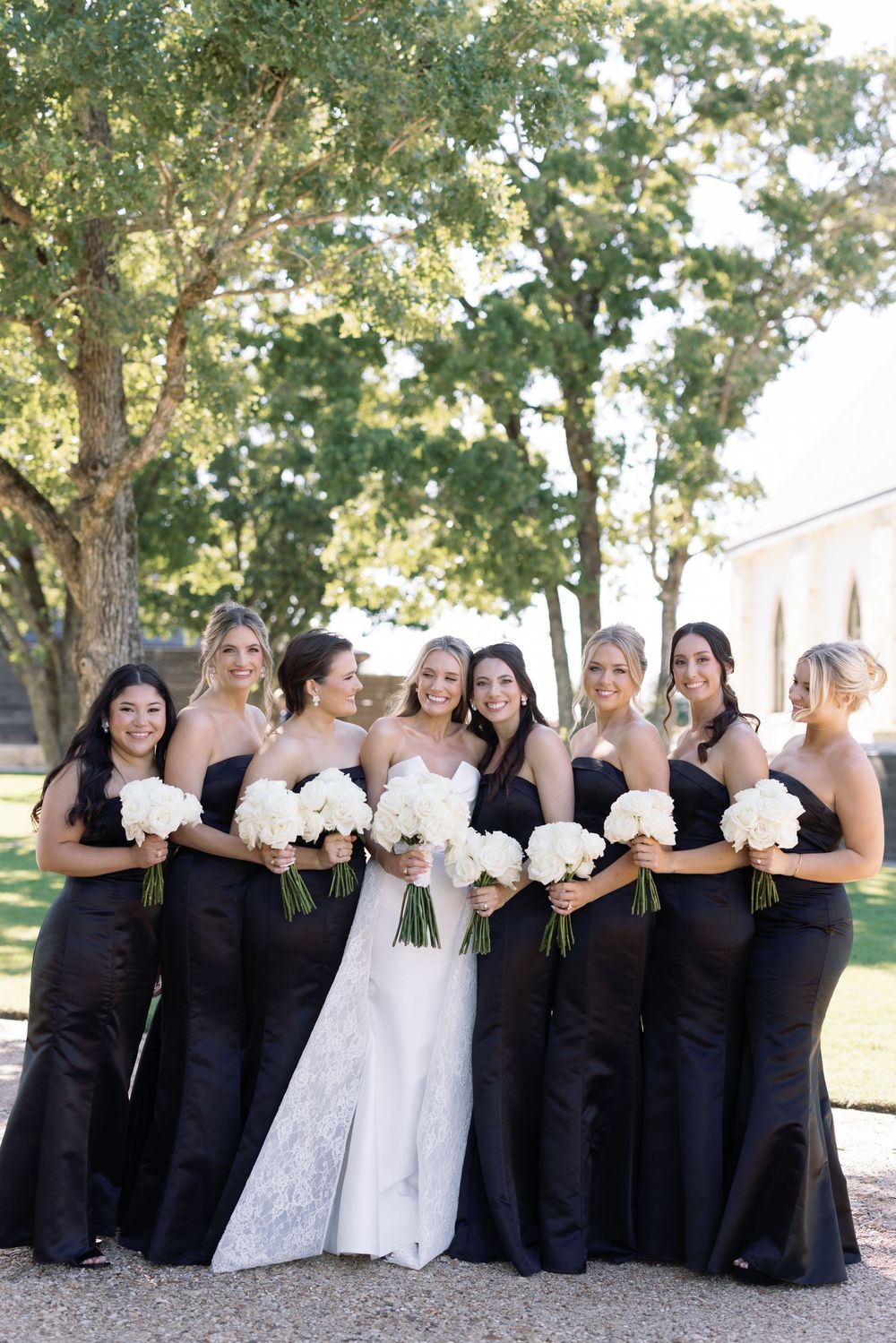 Classic wedding party group photo featuring bridesmaids in elegant black gowns holding white bouquets under shady trees.