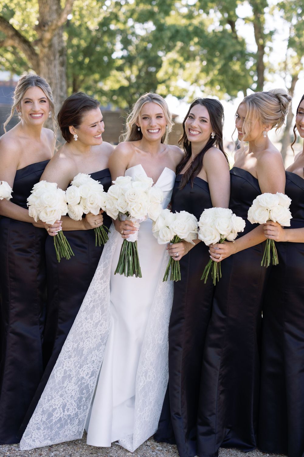 Wedding party stands together outdoors holding white bouquets with bride in lace gown and bridesmaids in elegant black dresses.