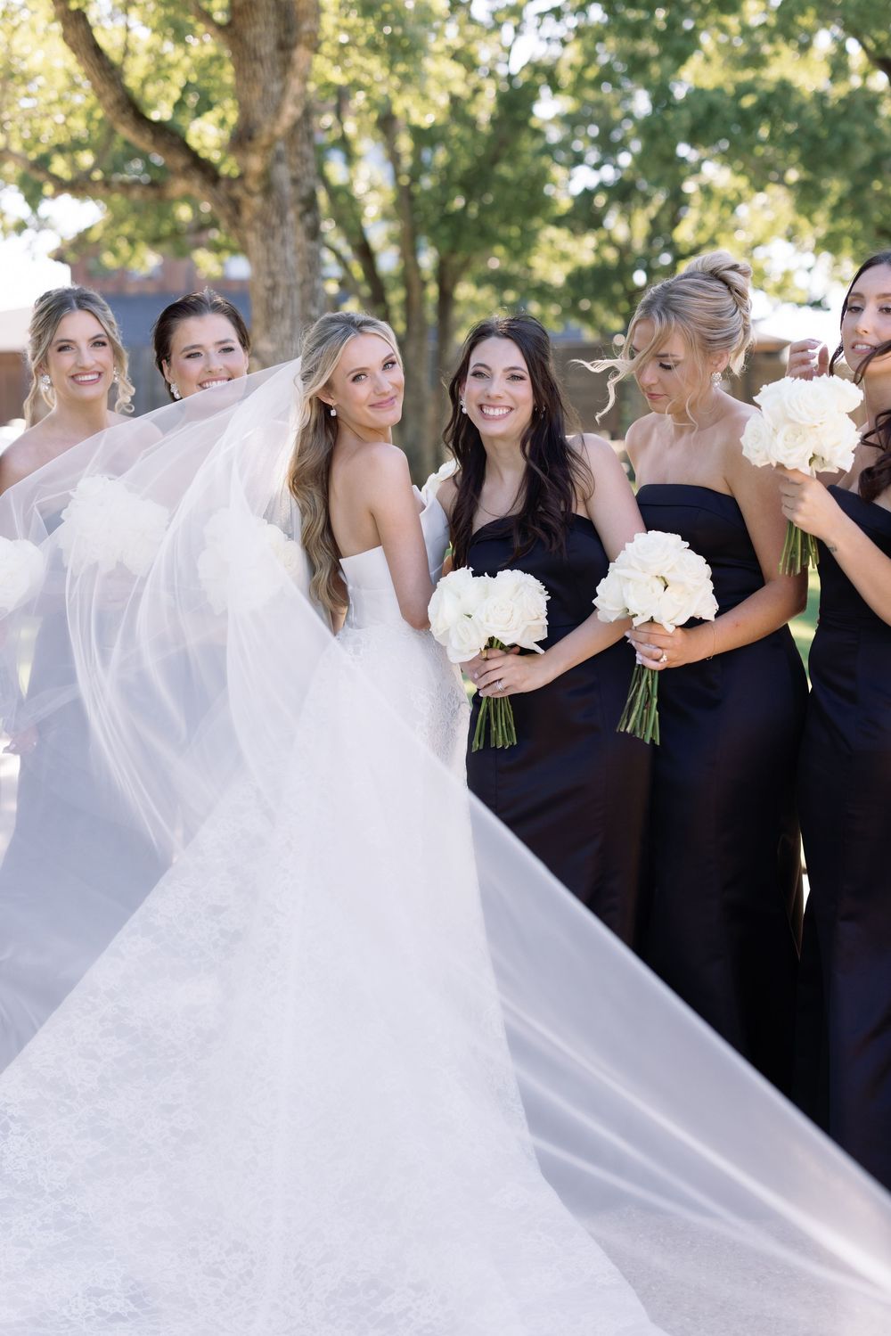 A group of bridesmaids in black dresses stand together with the bride in a flowing white gown and veil at an outdoor wedding.
