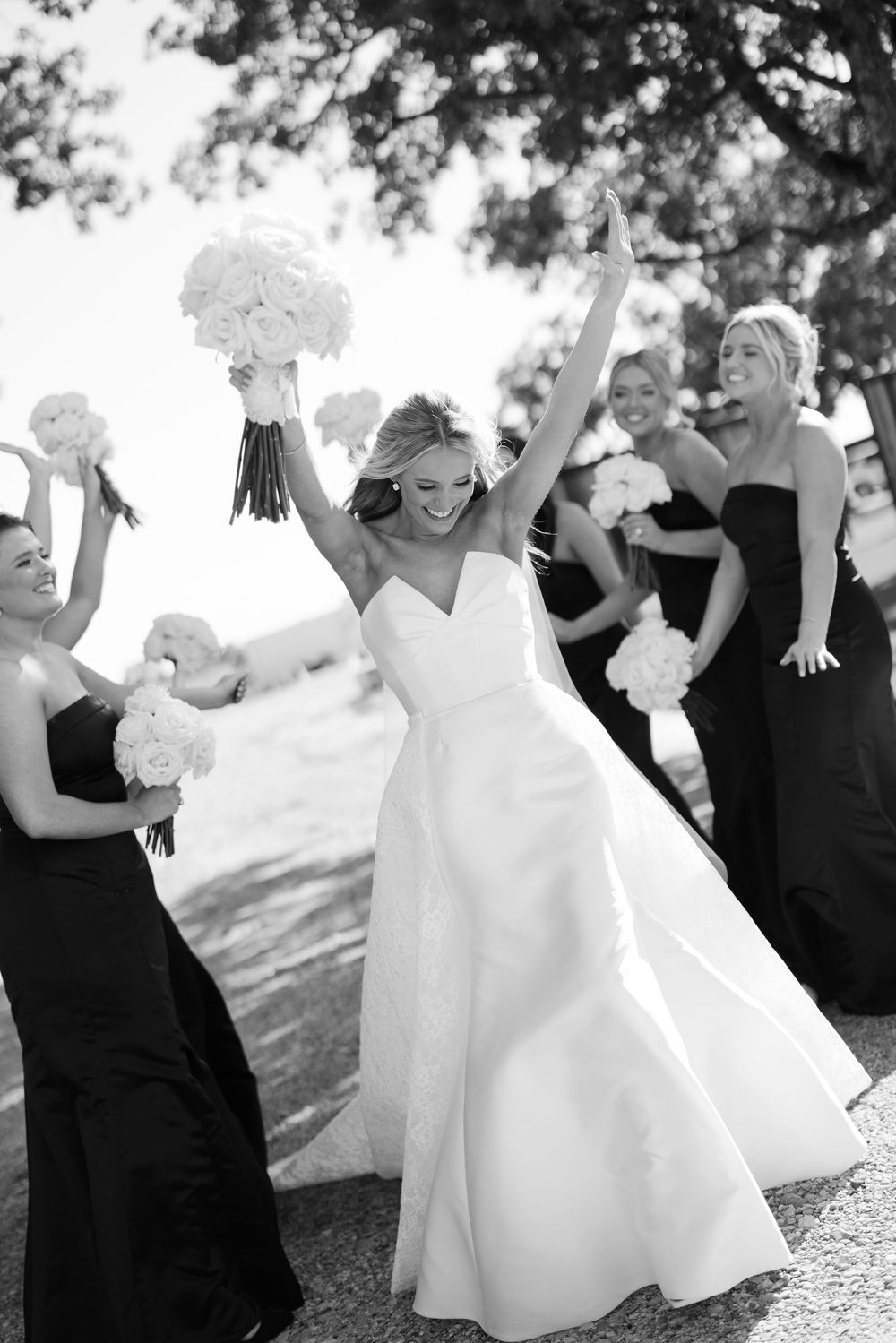 A bride in a white wedding gown celebrates joyfully with bridesmaids in black dresses during an outdoor ceremony.