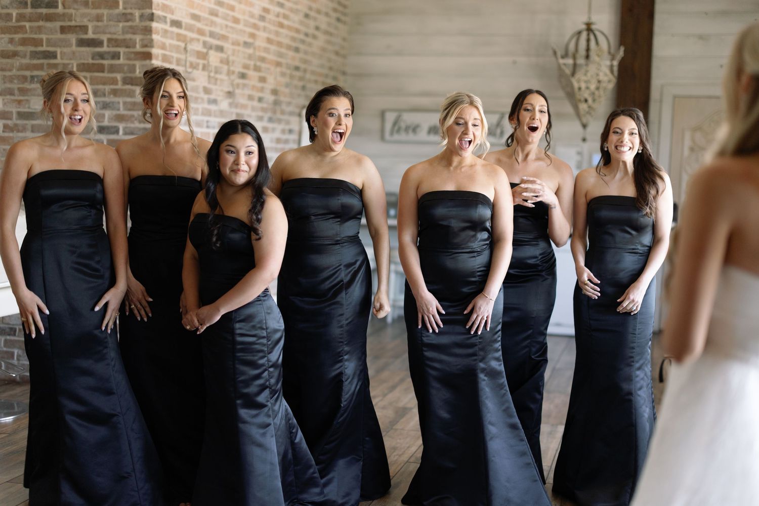 A row of bridesmaids wearing elegant strapless navy blue floor-length dresses stand together smiling in a brick room.