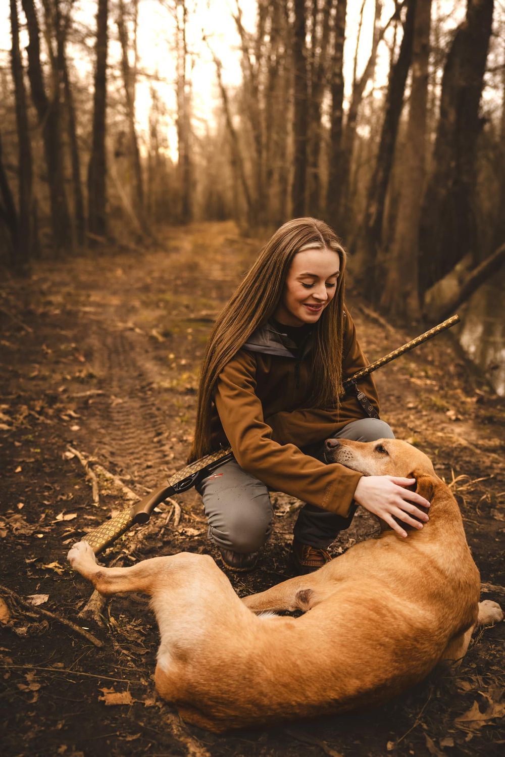 Person in brown jacket petting golden retriever on forest trail at sunset.