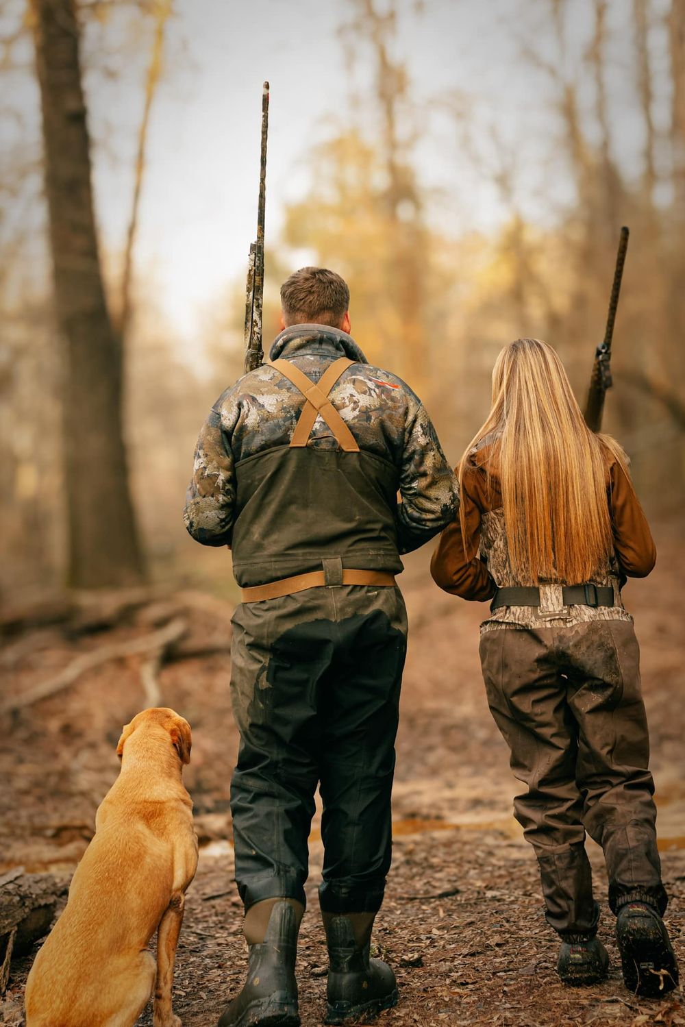 Two hunters in camouflage gear with rifles and a yellow dog looking into the autumn forest.