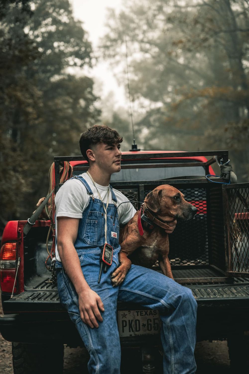 Young hunter in overalls sits on pickup truck bed with hunting dog.