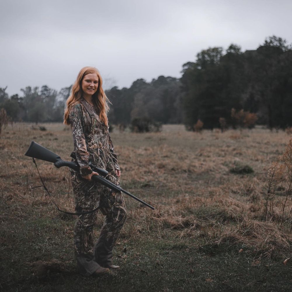 Hunter in camouflage gear poses with rifle in autumn field.