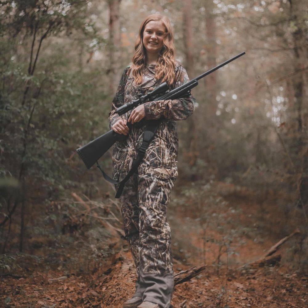 Hunter wearing camo clothing stands with rifle in autumn forest.