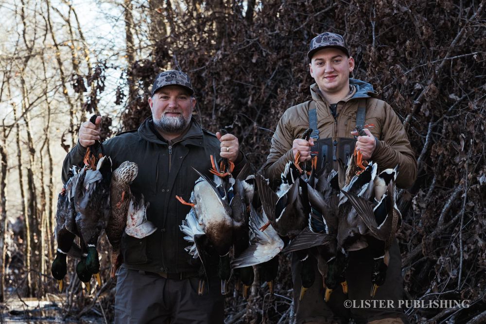 Two hunters display their waterfowl catch in a winter forest setting.