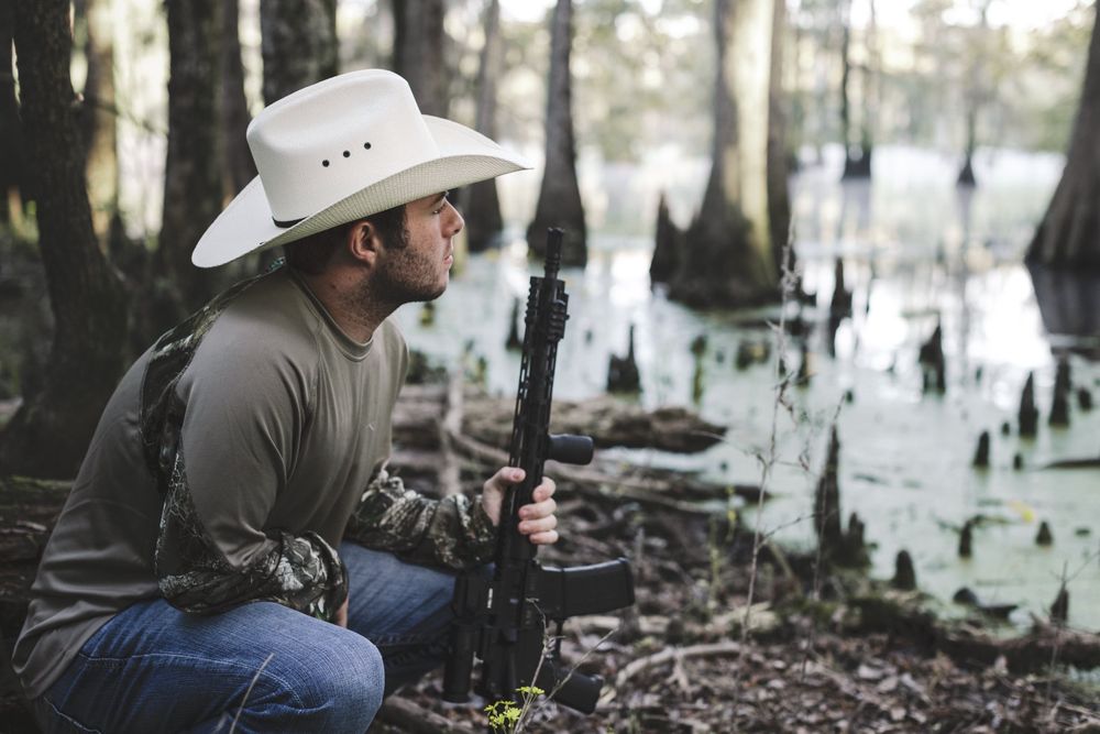 Hunter in cowboy hat crouches in swampy cypress forest with hunting gear.