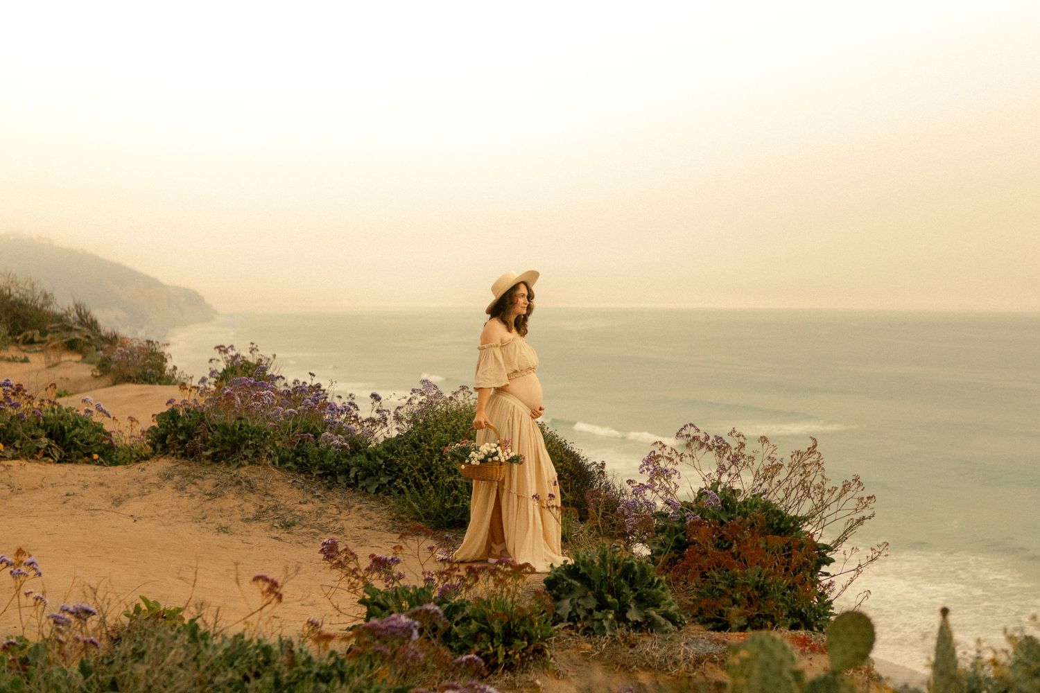 Person in flowing dress walks along coastal cliff edge with ocean views at sunset creating a dreamy silhouette.