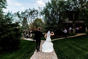 A couple walks away on a stone pathway surrounded by green trees and guests on their outdoor wedding day.