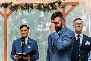 A group of groomsmen in navy suits standing by a wooden pergola decorated with greenery wait for the ceremony.