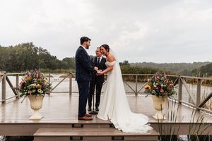 A wedding ceremony takes place on a wooden deck overlooking natural scenery with floral arrangements in urns.