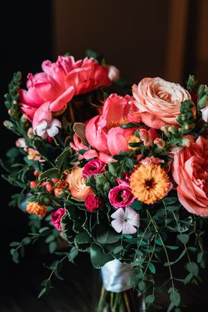 A vibrant floral arrangement with coral peonies, peachy roses, orange gerbera daisies and greenery in a moody darkened setting.
