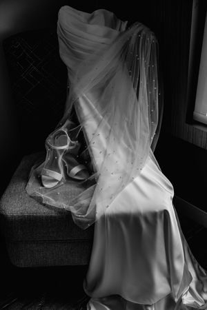 A black and white photograph of an elegant bridal gown and shoes arranged artistically on a chair.