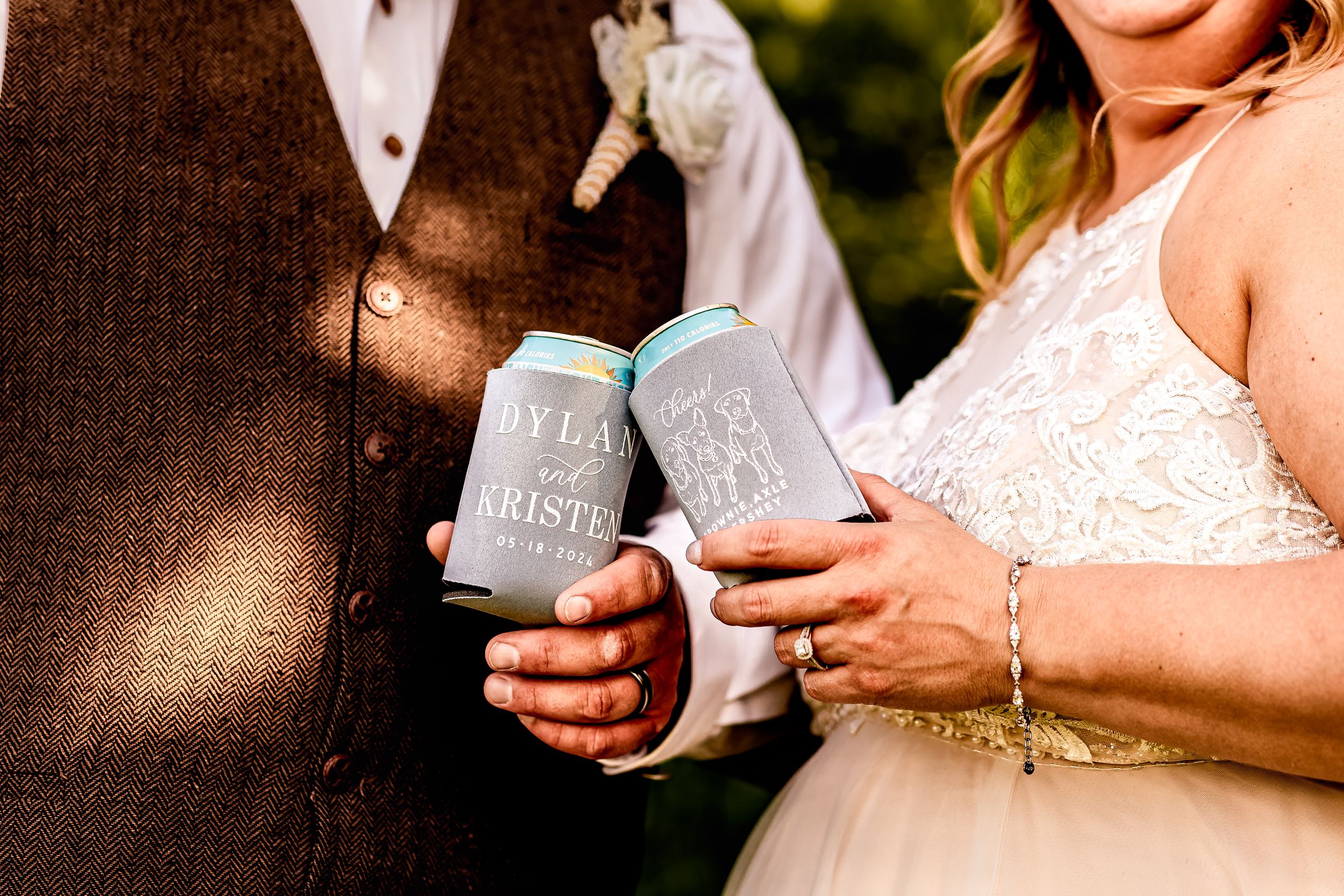 Bride and groom holding matching personalized grey koozies at a wedding celebration outdoors.