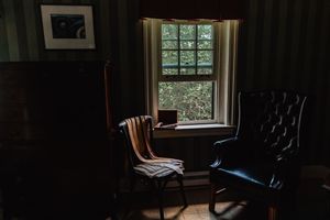 Moody interior scene with two contrasting chairs beside a bright window in a dark room with wooden paneling.