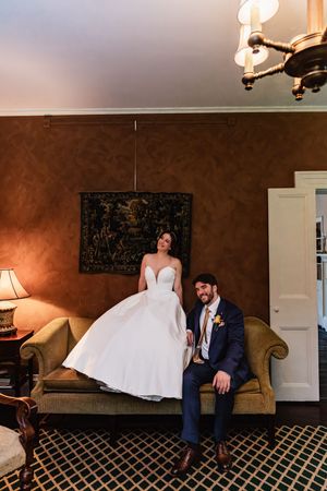 A couple in wedding attire share a tender moment on a brown chaise lounge with geometric patterned carpet below.