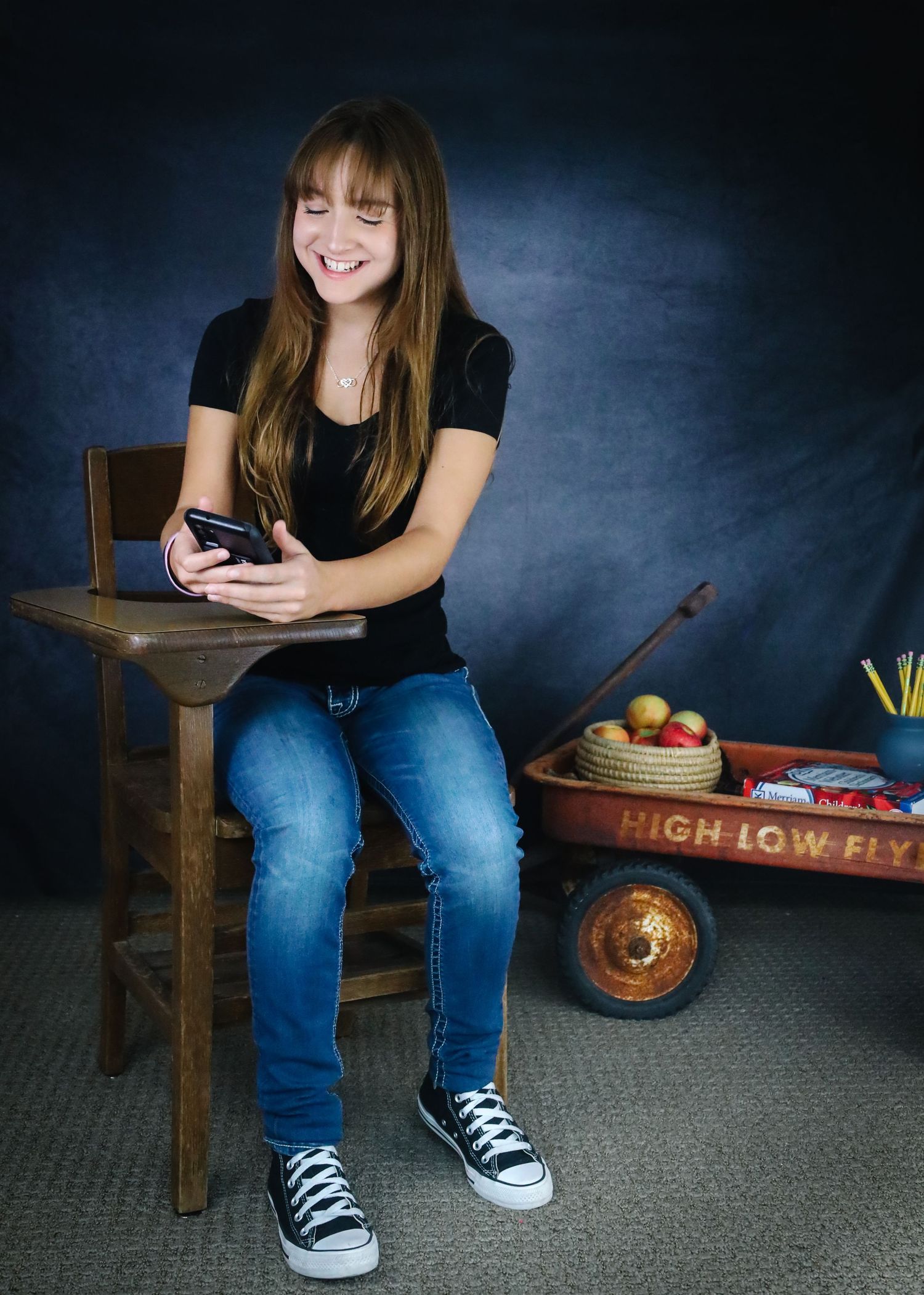 A casual portrait against a dark background with a wooden chair, red wagon, and person looking at their phone.