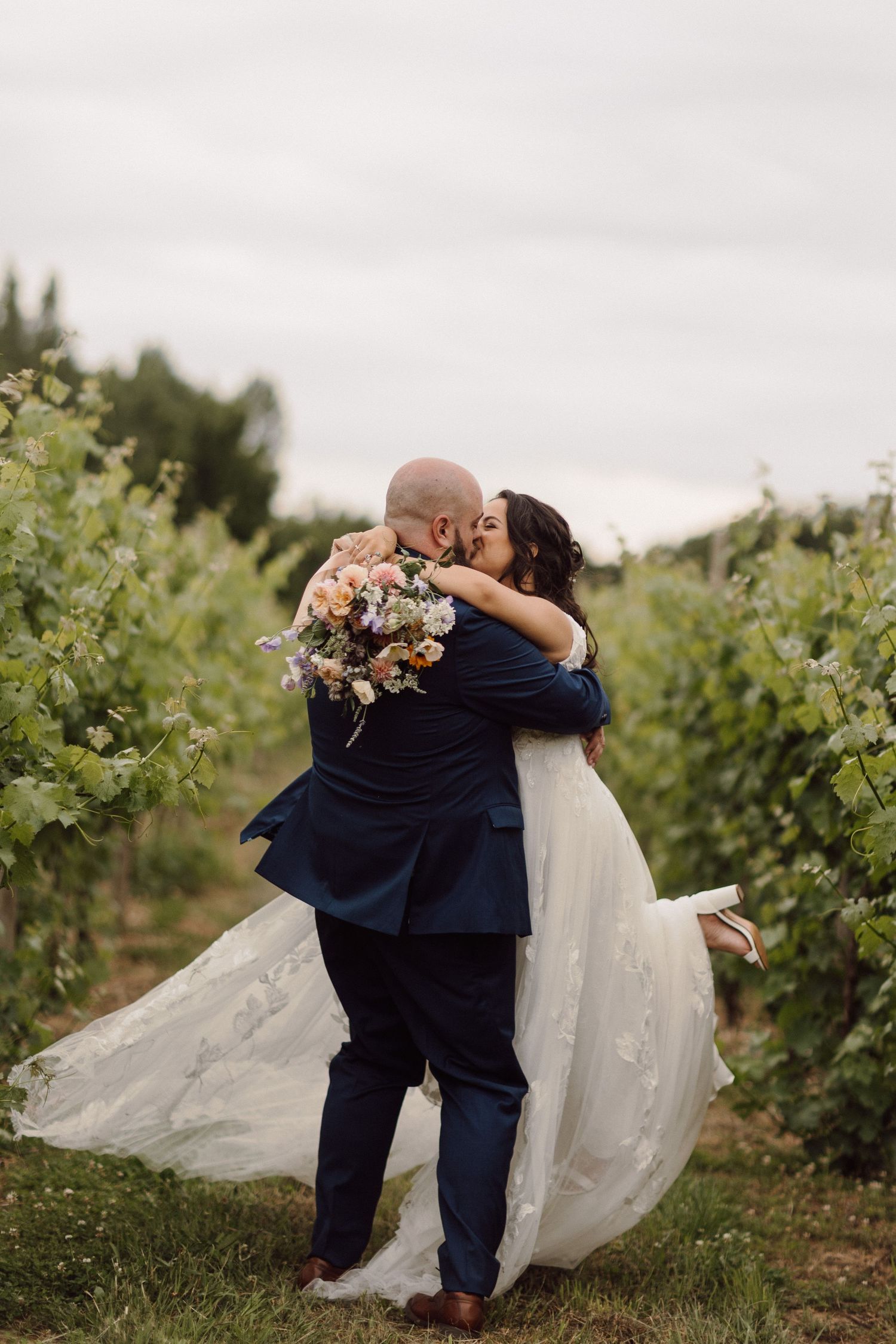 Romantic wedding couple embracing in a vineyard with flowing veil and floral bouquet.