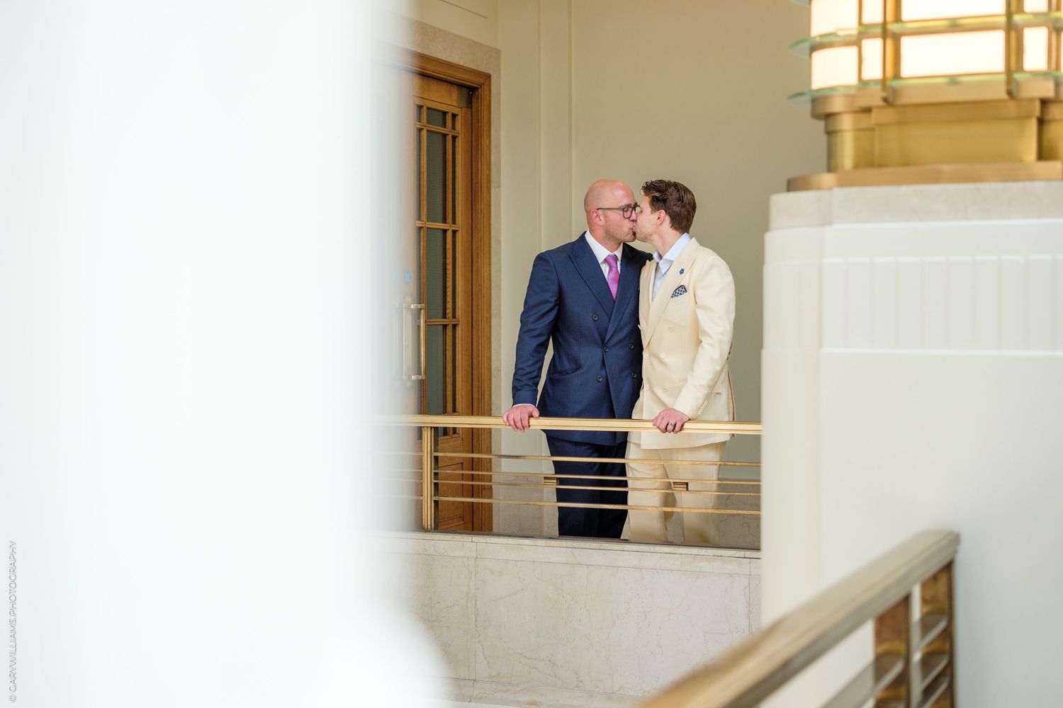 Two people share an intimate moment on a balcony with white railings and gold lighting fixtures.