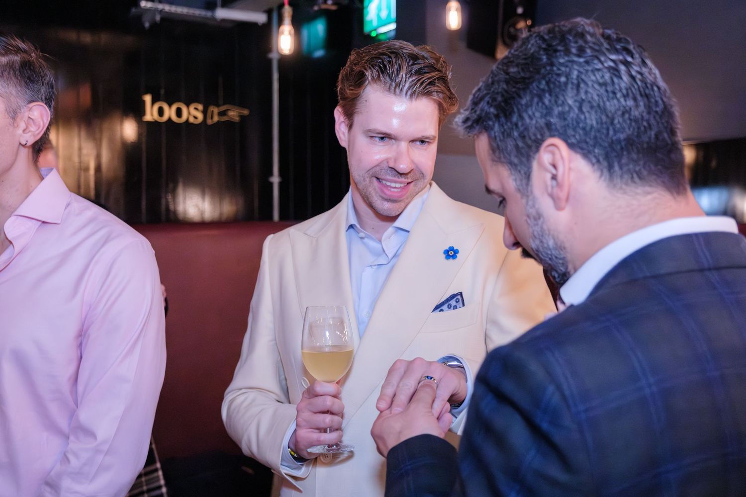 People having a social gathering at a bar, with one person holding a wine glass and wearing a light colored suit with a pocket square.