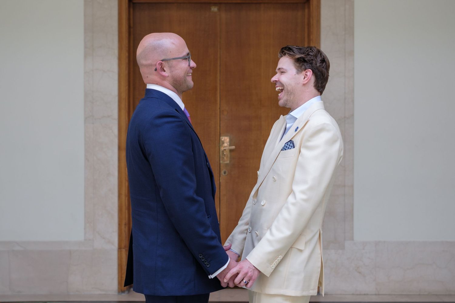 Two people in formal attire having an animated conversation in an office hallway near a wooden door.